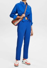 Modrá košile s knoflíky uvázaná v pase, odpovídající modré elegantní kalhoty, barevná tkaná kabelka a bílé tenisky s červenými akcenty.
