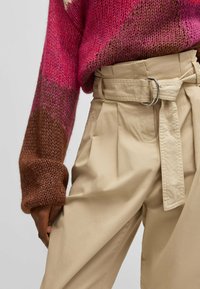 Strikket sweater i pink og brune nuancer med en struktureret overflade, parret med beige højtaljede bukser med bælte i taljen og plisseringer.