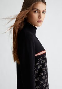 Maglione a collo alto nero con motivo decorativo sul petto e logo, caratterizzato da una striscia rossa. Tessuto morbido e vestibilità rilassata.