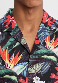 Camisa negra de manga corta con un vibrante estampado floral tropical, que presenta tonos rojos, naranjas y verdes, y un cuello con muesca y un cierre de botón.