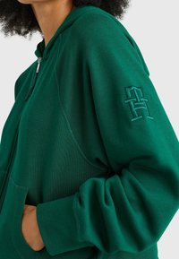 Haut à capuche zippé vert avec une poche frontale, des poignets côtelés et un logo brodé sur la manche. Tissu doux avec une texture lisse.