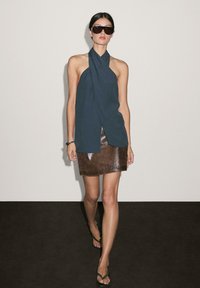 Top halter blu scuro abbinato a una mini gonna in pelle marrone. La modella indossa occhiali da sole neri oversize e sandali. Texture lisce, senza motivi.