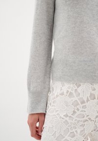 Pulover tricotat gri deschis cu manșete încrețite, asociat cu o fustă albă din dantelă, care prezintă modele florale și elemente transparente.