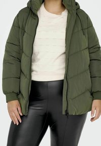 Doudoune vert olive avec capuche, dotée d'une fermeture éclair à l'avant et de poignets élastiques. Portée par-dessus un pull crème et un legging en simili cuir noir.