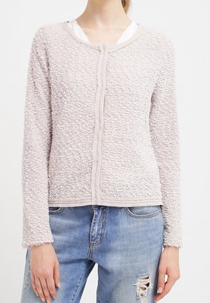 Femme portant un cardigan texturé rose clair sur un haut blanc assorti à un jean bleu clair déchiré, vue du cou aux hanches.