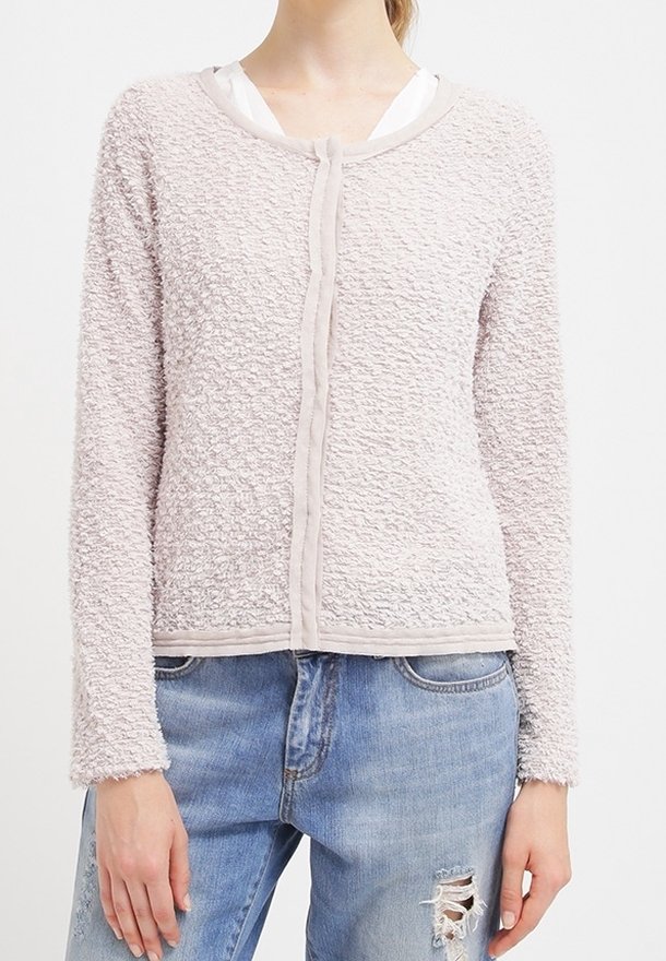 Femme portant un cardigan texturé rose clair sur un haut blanc assorti à un jean bleu clair déchiré, vue du cou aux hanches.