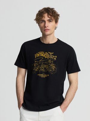 Giovane uomo con capelli ricci che indossa una maglietta nera con grafica vintage di una motocicletta gialla e testo, abbinata a pantaloni bianchi, mani nelle tasche.