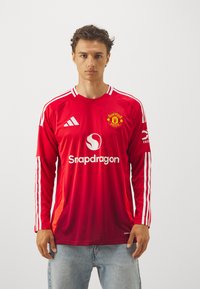 Raudonas ilgomis rankovėmis džemperis su baltais Adidas dryžiais, apvaliu kaklu, Manchester United logotipu ir "Snapdragon" prekių ženklu priekyje.