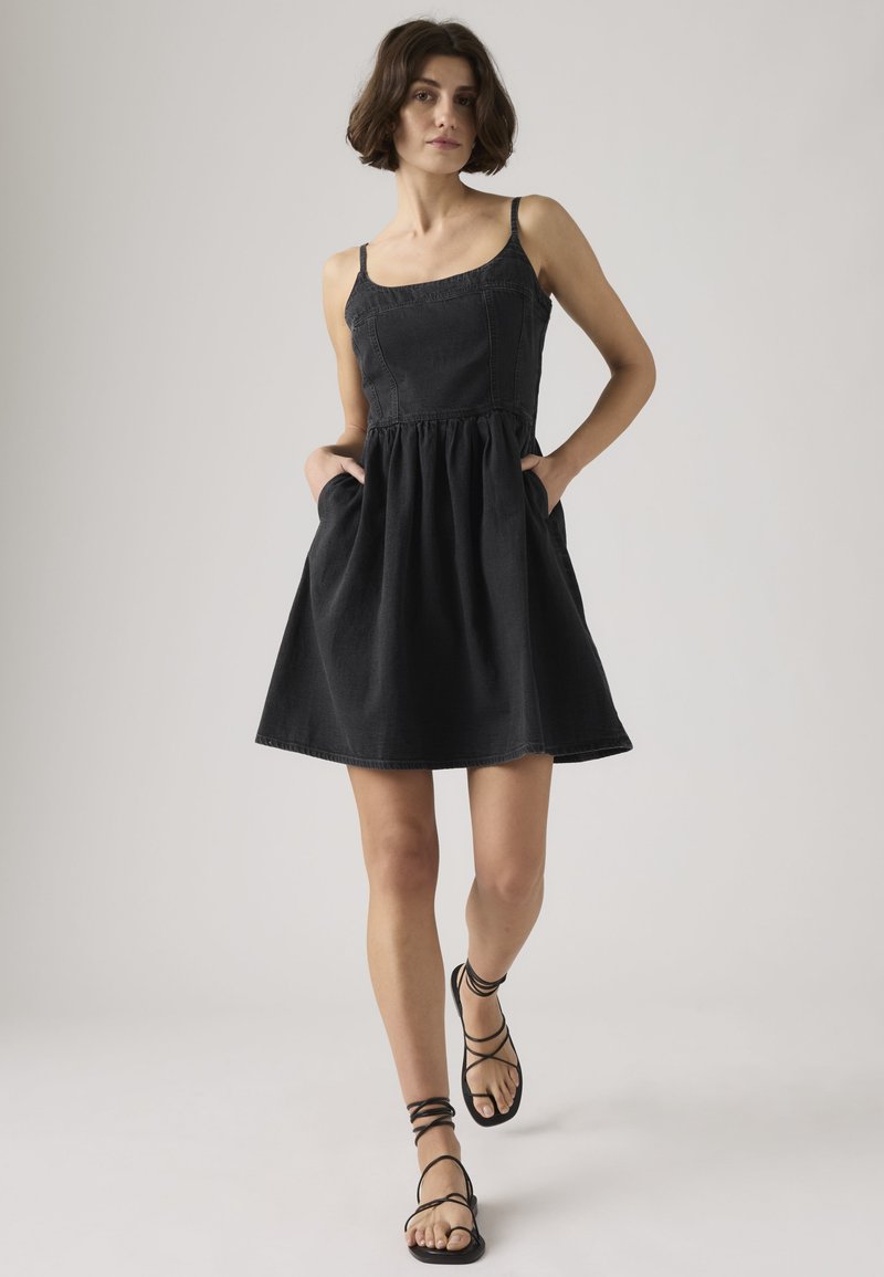 Vrouw met kort krullend haar, gekleed in een mouwloze zwarte jurk met zakken en zwarte sandalen met bandjes, staand tegen een effen achtergrond.