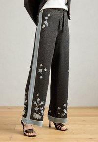Pantalons larges noirs avec une texture brillante, ornés d'une broderie florale bleue et argent le long des côtés et d'un poignet à motifs.