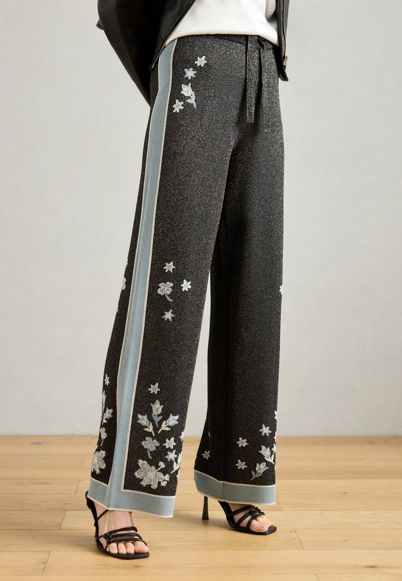 Pantalons larges noirs avec une texture brillante, ornés d'une broderie florale bleue et argent le long des côtés et d'un poignet à motifs.