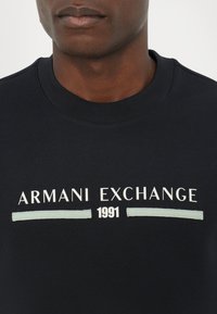 Mørkeblå crewneck sweatshirt lavet af bomuld, med hvid og lyseblå grafisk tekst: "ARMANI EXCHANGE" og "1991." Enkelt design, ingen mønstre.