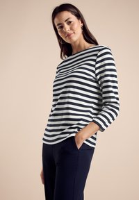 Pruhovaný bavlněný top s třičtvrtečními rukávy v námořnické modré a bílé barvě. Má lodičkový výstřih a volný střih, kombinovaný s tmavě námořnickými kalhotami.