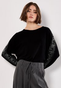 Maglione nero, comodo e largo, con maniche ampie decorate da embellishments in argento. Abbinato a pantaloni grigi a righe sottili e una silhouette rilassata.