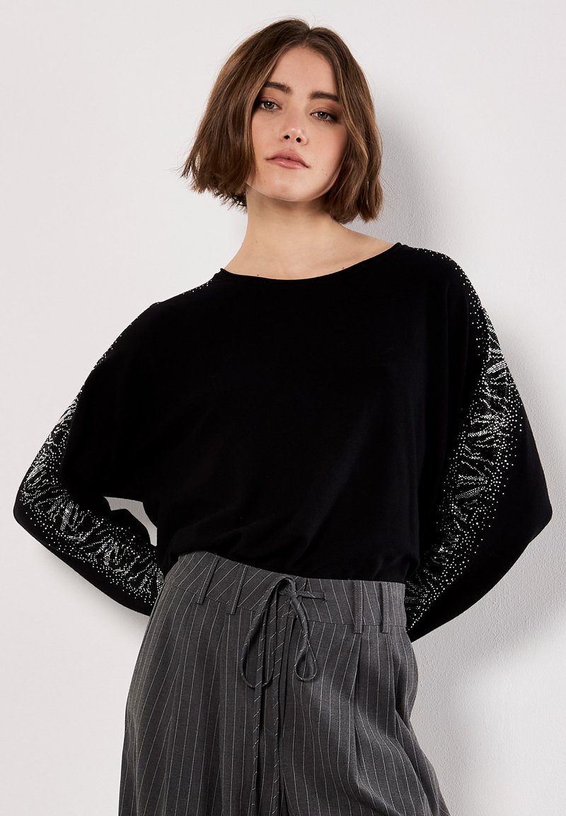 Pull noir ample avec des manches larges orné d'ornements en argent. Associé à un pantalon à rayures grises et une silhouette décontractée.