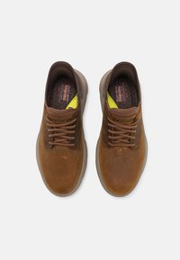 Braune Ledersneaker mit dunkelbraunen Schnürsenkeln, strukturierten braunen Akzenten und gelben Details auf der Innensohle. Abgerundetes Design mit gummierten Außensohlen.