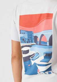Camiseta de algodón con un gráfico vibrante de barcos y edificios, en colores predominantemente azul y coral, con una impresión texturizada.