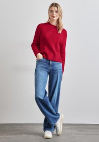 Maglione rosso a coste con scollo rotondo, abbinato a jeans blu a gamba larga e scarpe da ginnastica bianche. Texture liscia, vestibilità casual e stile rilassato.