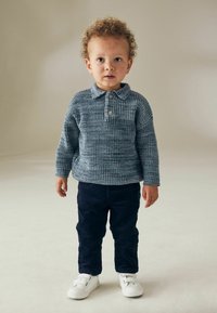 Bambino che indossa un maglione lavorato a maglia blu con colletto e bottone, abbinato a pantaloni in velluto a coste blu navy e sneakers bianche.