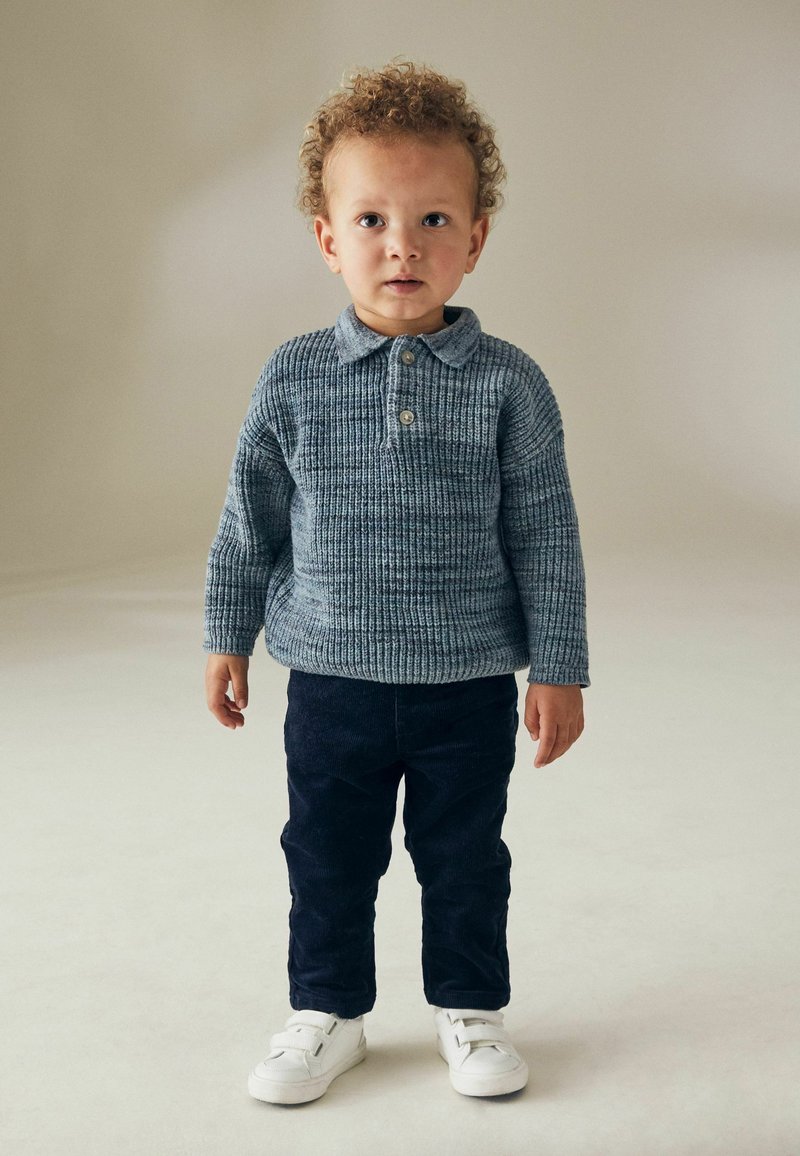 Bambino che indossa un maglione lavorato a maglia blu con colletto e bottone, abbinato a pantaloni in velluto a coste blu navy e sneakers bianche.