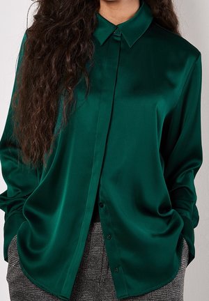 Osoba ubrana w ciemnozieloną satynową bluzkę z długim rękawem, wsuniętą w szare, wysokowzsokie spodnie w kratę, ręce w kieszeniach, z długimi kręconymi włosami.