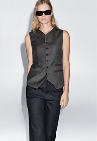 Charcoal gray sleeveless vest with a notched hem, featuring a five-button front closure and two side pockets, paired with dark denim trousers.