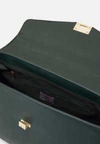 Zielona syntetyczna torebka z złotym zapięciem, z wewnętrzną kieszenią na zamek i etykietą z materiału. W podszewce, z gładkimi i teksturowanymi detalami.