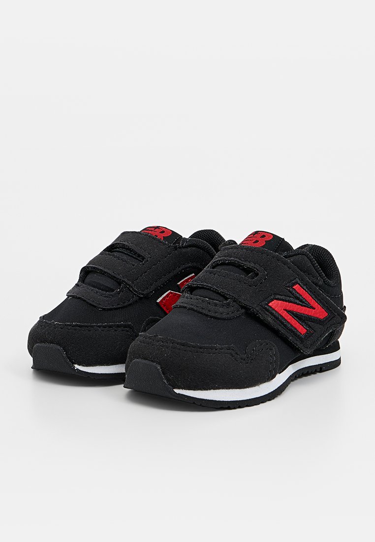 Chaussures de sport noires avec une sangle Velcro, logo 'N' rouge contrastant et semelle blanche avec des accents noirs, fabriquées en matériau durable.