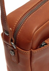 Borsa in pelle marrone con una texture liscia, dotata di una cerniera argentata e dettagli in metallo neri. Cuciture visibili lungo i bordi. Forma compatta.