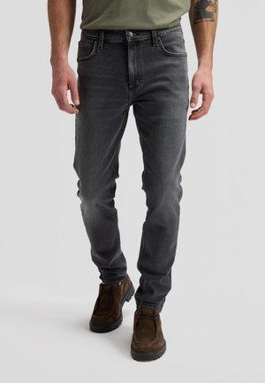 Mand iført slidte sorte slim fit-jeans, olivengrøn t-shirt og brune ruskindssnørestøvler, stående mod en ensfarvet baggrund.