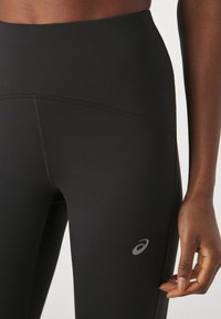 High-waisted black leggings made of stretchy fabric. Features a smooth texture and a small reflective logo on the left side.