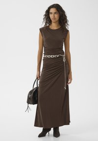 Femme aux cheveux bouclés portant une robe marron sans manches froncée avec une ceinture chaîne argentée, des bottines noires, et tenant un sac à main noir.