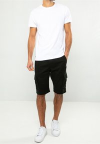 Camiseta de algodón blanca combinada con pantalones cortos de carga negros, con bolsillos laterales, llevados con zapatillas blancas. Outfit simple y casual.
