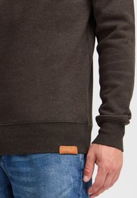 Felpa marrone scuro con finitura testurizzata, polsini e orlo a coste. Presenta un'etichetta in pelle color beige vicino al fondo. Indossata con jeans blu.
