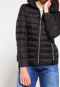 Veste rembourrée noire avec fermeture éclair, dotée d'une capuche et de coutures diagonales. Portée sur un haut rayé et associée à un jean bleu.