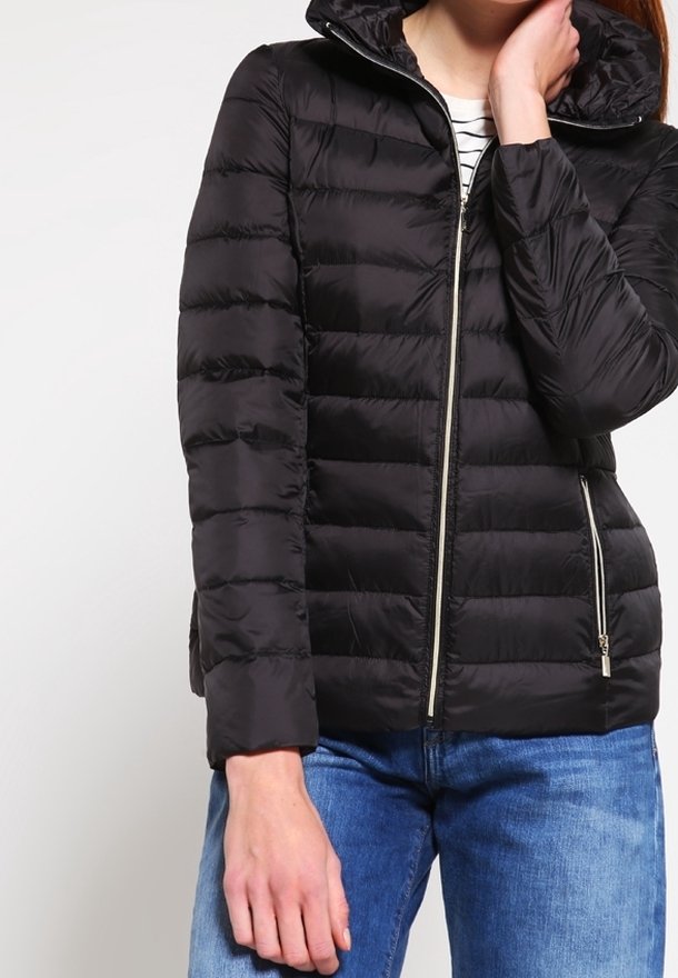 Veste rembourrée noire avec fermeture éclair, dotée d'une capuche et de coutures diagonales. Portée sur un haut rayé et associée à un jean bleu.