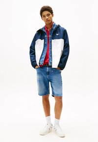 Chaqueta cortavientos azul marino y blanca con acentos de mezclilla, camiseta roja y pantalones cortos de mezclilla. Zapatillas blancas y calcetines estampados completan el conjunto.