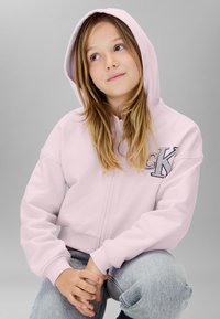 Sudadera rosa con cremallera y una gran letra "K" bordada en azul. Cuenta con capucha con cordón y puños acanalados, combinada con jeans grises.