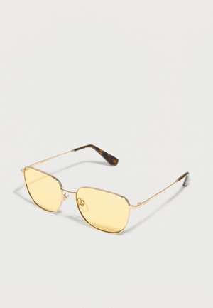 Solbriller med gullramme, gule linser og straumønstrede tempelender på hvit bakgrunn.