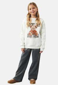 Biały sweatshirt z grafiką niedźwiedzia, napisem, z prążkowanymi mankietami; w połączeniu z ciemnoniebieskimi spodniami o szerokich nogawkach i brązowymi trampkami z beżowymi akcentami.