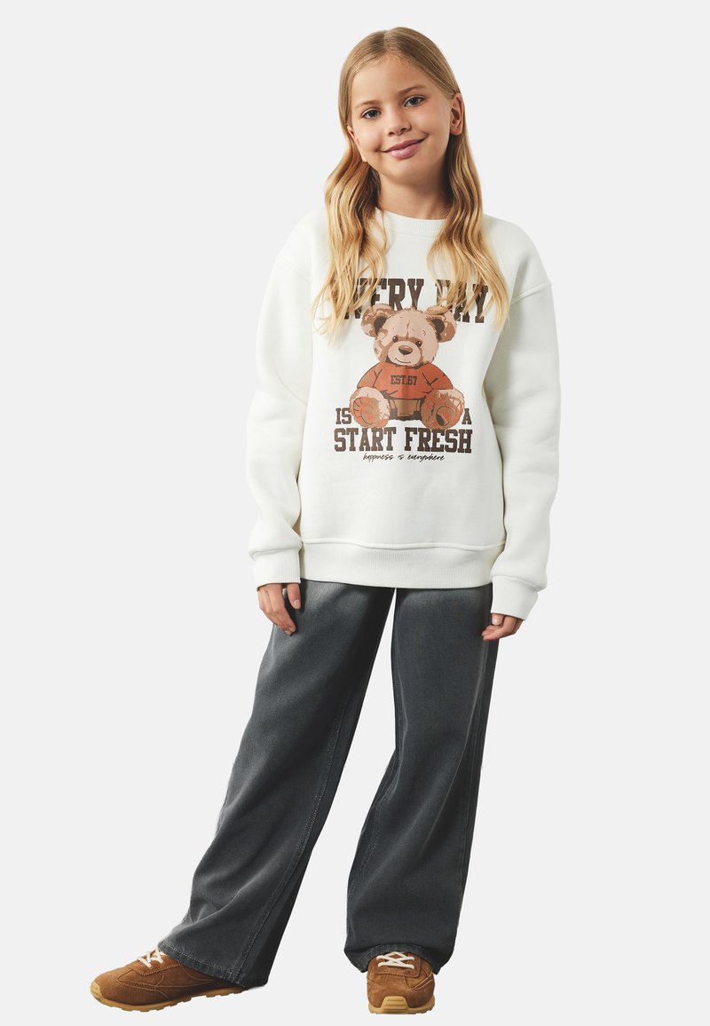 Biały sweatshirt z grafiką niedźwiedzia, napisem, z prążkowanymi mankietami; w połączeniu z ciemnoniebieskimi spodniami o szerokich nogawkach i brązowymi trampkami z beżowymi akcentami.