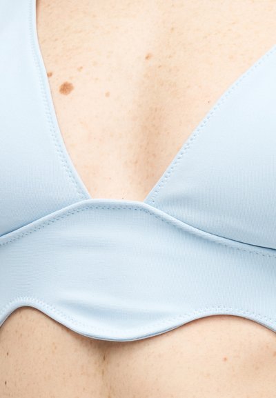 Κοντινό πλάνο ανοιχτόχρωμου μπλε τοπ μπικίνι που φοριέται πάνω σε δέρμα με φακίδες, με μια ορατή καφέ ελιά κοντά στην άνω άκρη του ενδύματος.