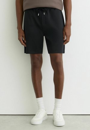 Mężczyzna w czarnych szortach ze ściągaczem, białych skarpetkach i białych trampkach stojący na tle jasnej ściany.