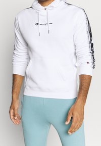 Weißer Hoodie aus Baumwollmischung mit einer Vorderentasche, verstellbarem Kordelzug und schwarzen Logo-Akzenten an den Ärmeln.