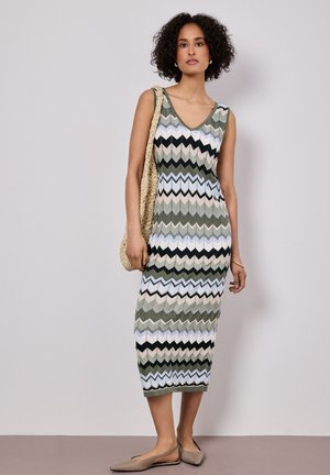 Nainen, joka pukeutunut hihattomaan, vihreän, mustan, beigen ja valkoisen zickzack-kuvioiseen midimekkoon, pitää käsissään kudottua olkalaukkua ja seisoo yksiväristä seinää vasten.