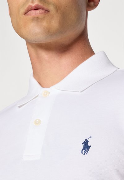 Valkoinen polo-paita, jossa on kolme nappia ja tummansininen brodeerattu logo vasemmalla puolella, sekä teksturoitu kangas.