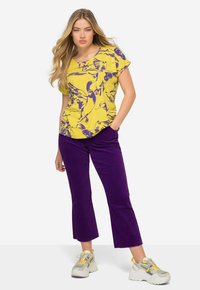 Gul kortärmad blus med lila fågeltryck, kombinerad med lila utsvängda byxor och kraftiga grå sneakers med gula accenter.