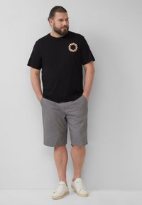 Zwarte katoenen t-shirt met een cirkelvormige oranje graphic op de borst, gecombineerd met grijze shorts en witte sneakers. Casual outfit, ontspannen pasvorm.