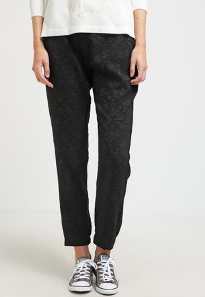 Person wearing black textured floral pants and grey lace-up sneakers, standing against a plain white background.