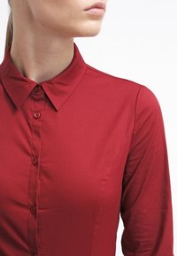 Chemise rouge à boutons avec un col classique et des manches longues. Tissu lisse et légèrement brillant ; boutons assortis sur le devant.